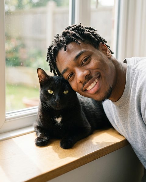 A man smiling cheek to cheek with his black cat on a window sill, a portrait made by Stay
